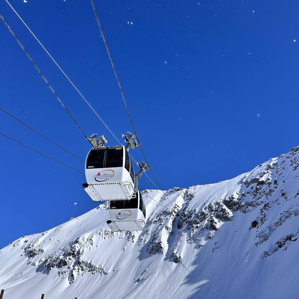 Two gondola cabins climbing towards the Pic Blanc against a deep blue sky, with a snow-covered ridge below.