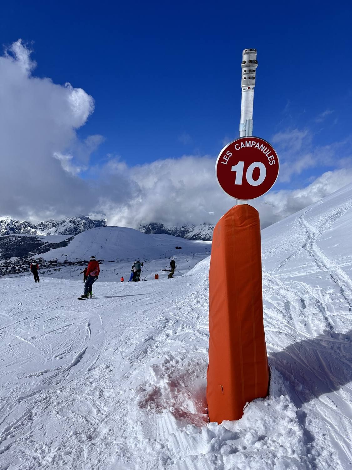 An orange padded piste marker carrying a red sign reading Les Campanules 10, with skiers and the resort on the slope behind.