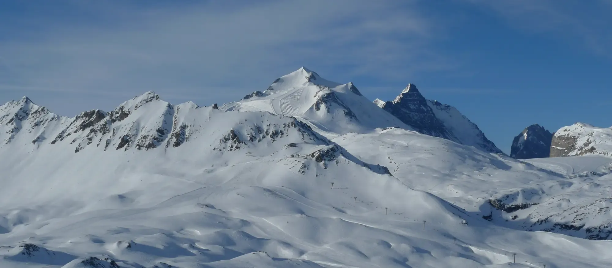 A wide panorama of snow-covered peaks and pistes above Tignes under a blue sky, with lift lines crossing the lower slopes.