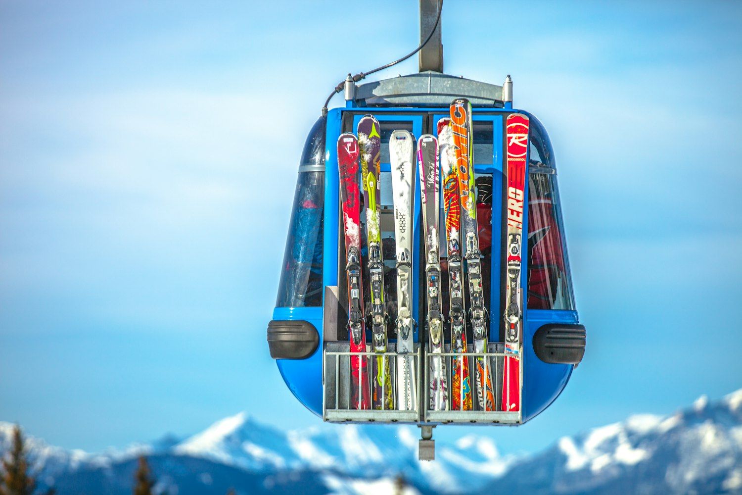 Close-up of a blue gondola cabin with colourful skis racked on its rear holder, snowy mountain peaks blurred in the background.