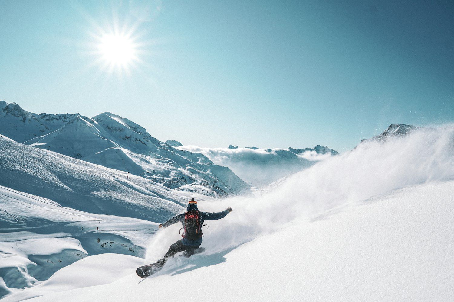 Snowboarder carving a deep turn through fresh powder in bright sunshine, spraying a cloud of snow, with snow-covered peaks and blue sky behind.
