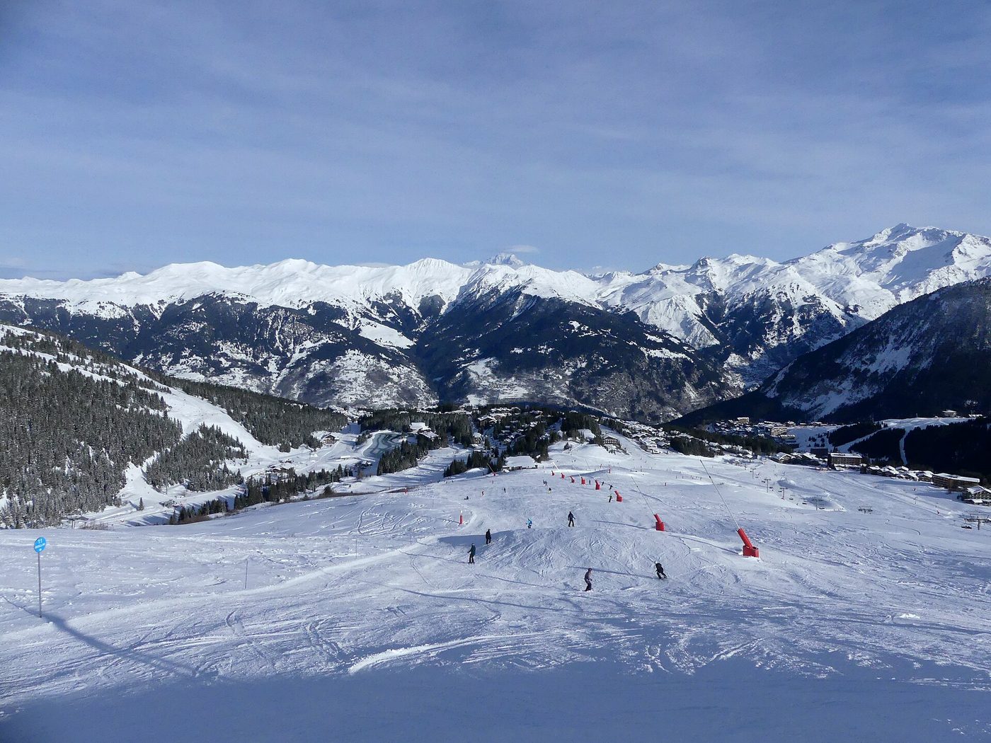 Skiers on a wide groomed piste descending through snowy terrain towards Courchevel 1850 with Alps peaks beyond