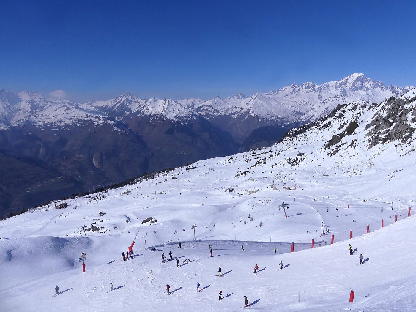 A wide groomed piste at Les Arcs with skiers on the snow and the Mont Blanc massif visible across the Tarentaise valley