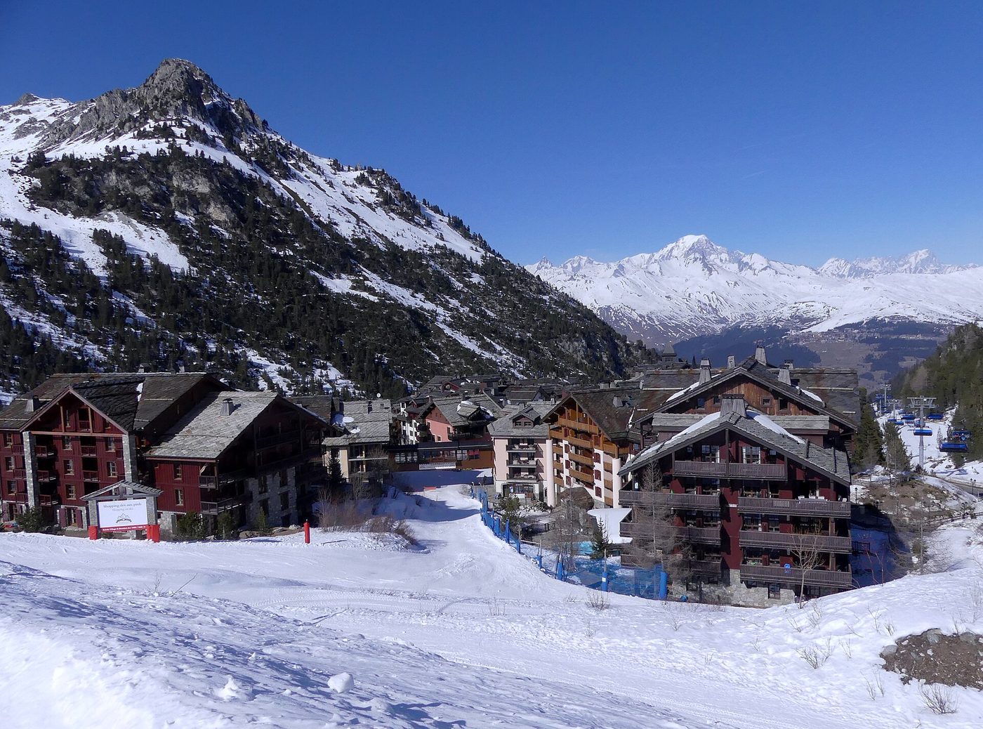 Snow-covered apartment buildings of Arc 1950 ski village in winter with a ski piste running past and mountains in the background