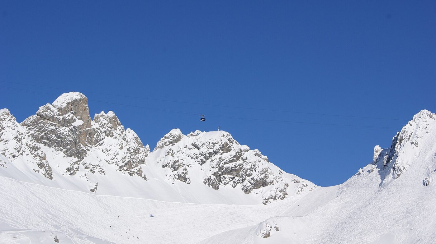 A cable car gondola crossing between dramatic rocky Arlberg mountain peaks covered in deep winter snow under a brilliant blue sky
