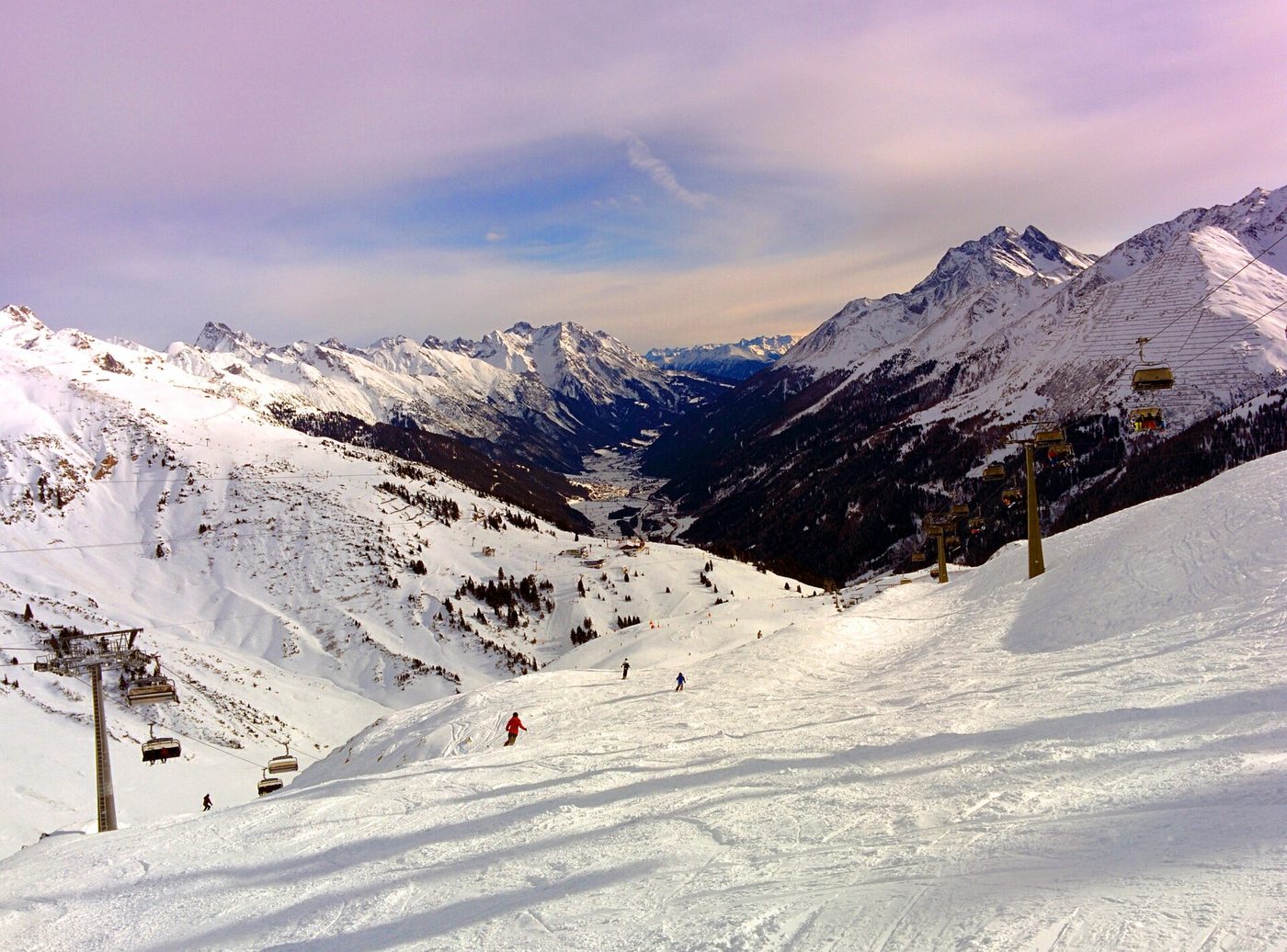 Wide view looking down ski pistes toward St. Anton am Arlberg in the valley below, with mountain ridges on either side and skiers on the slopes