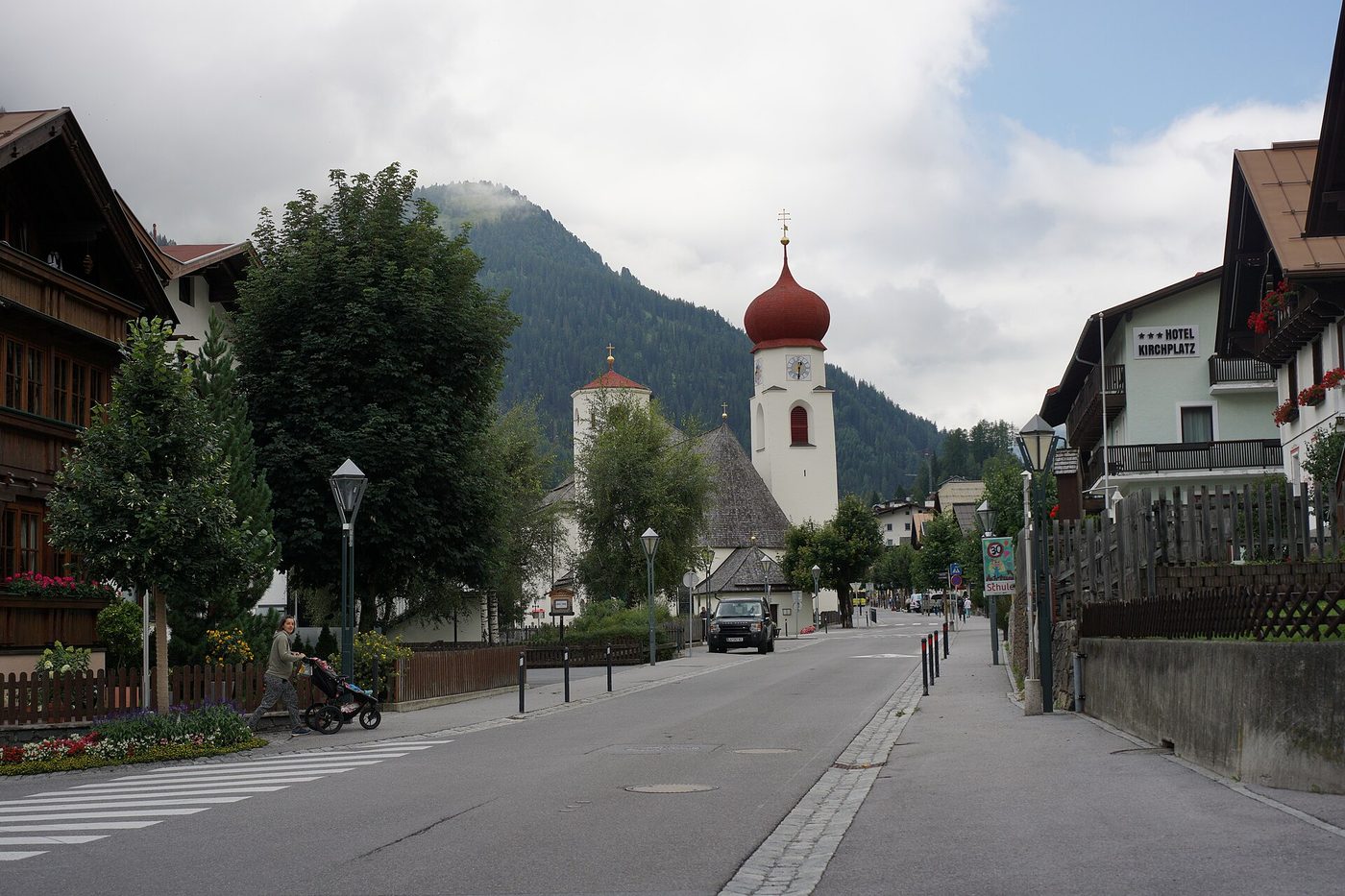 The main Dorfstrasse street through St. Anton am Arlberg in summer, with traditional Austrian buildings lining the road and the white parish church with its red onion dome in the background