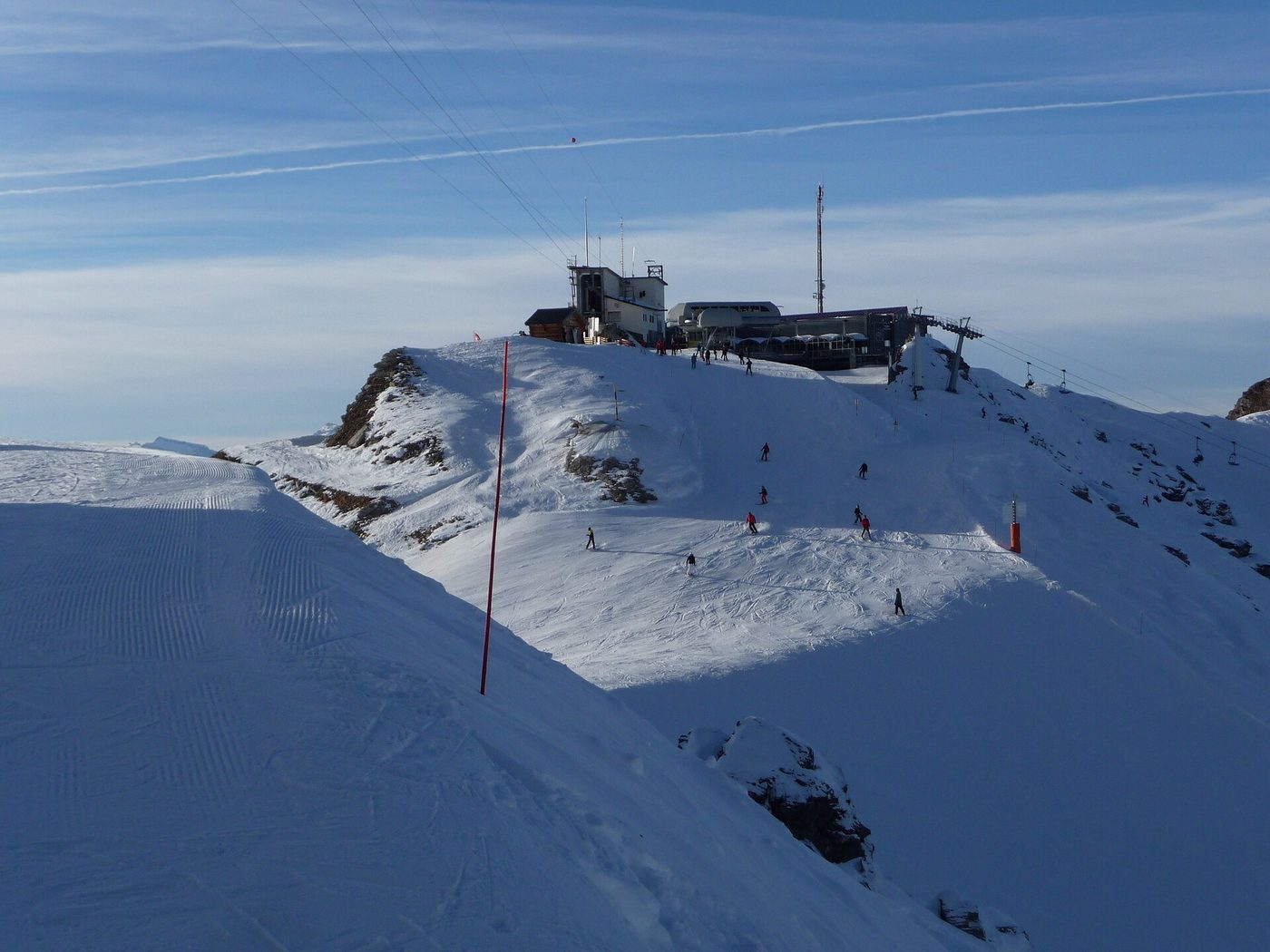 Skiers descending a groomed piste below the top station at Verbier's Mont-Fort with snow-covered terrain stretching away