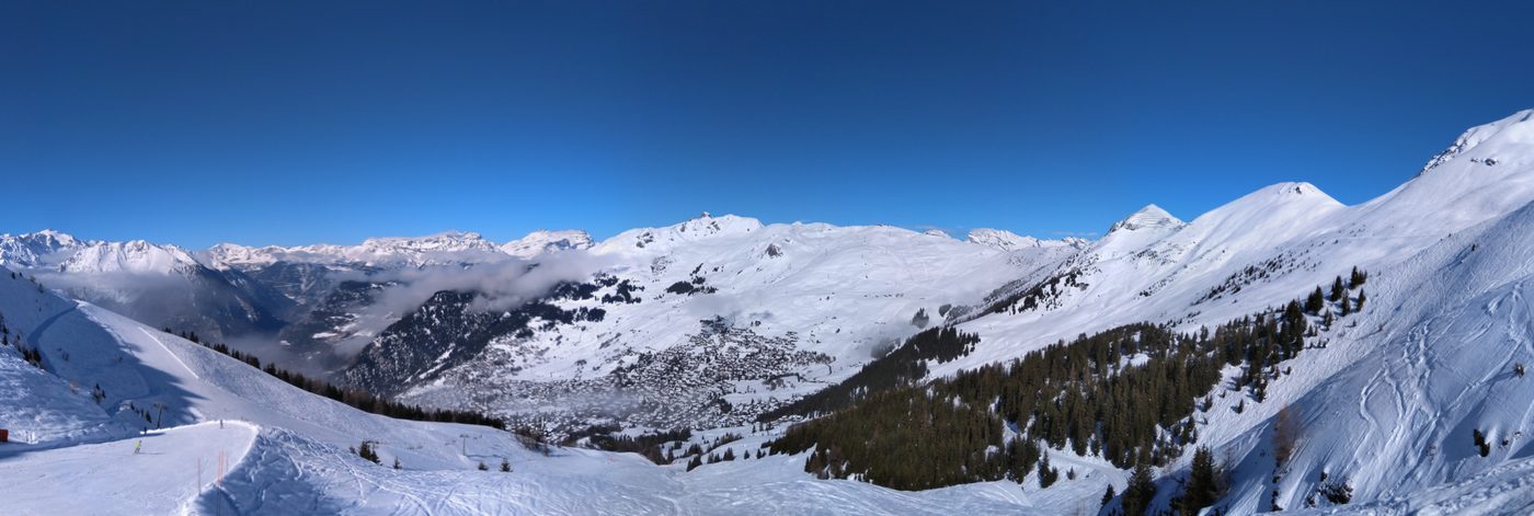 Panoramic view of Verbier village in its Alpine valley surrounded by snow-covered ski slopes and wooded hillsides under a blue winter sky